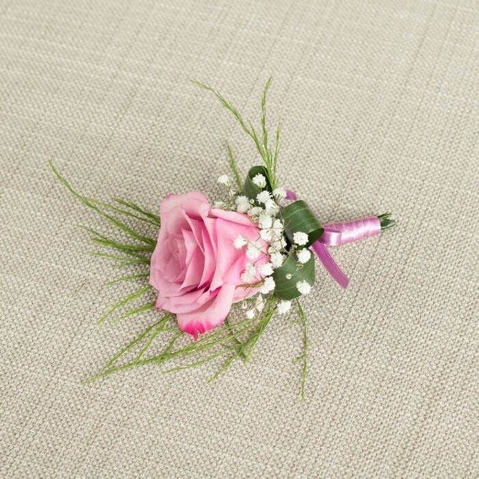 Set on a softly textured, light beige linen cloth, this pink rose boutonniere is captured from an overhead perspective, as if laid out on a dressing table in a Grove Park home while final preparations for a ceremony quietly unfold. The central rose is a richly layered beauty, its petals tightly clustered at the heart and gradually unfurling outward in a spiral of colour, transitioning from a gentle blush pink at the inner whorls to a slightly deeper, more saturated pink along the outer, softly ruffled edges. The velvety surface of the petals catches the ambient light, creating delicate highlights and shadows that emphasise the flower's romantic fullness. Encircling the base of the rose, clusters of tiny, cloud-like white baby's breath blossoms add a soft, airy texture, each miniature flower head contributing to a halo effect that lightens the design. Nestled beneath and behind the blooms are two complementary styles of greenery: fine, wispy blades of bright green foliage extend outwards in slender arcs, suggesting movement and a garden-fresh feel, while a larger, darker green leaf is carefully folded and wrapped around the lower stems, providing structure and a polished, tailored silhouette. The stems of the boutonniere are neatly bound with a slim, light pink satin ribbon that matches the rose's softer tones, the ribbon wrapped snugly and finished with a small, elegant bow whose short tails rest gently on the linen. The neutral background, with its visible woven texture, gives the photograph a natural, understated character that allows the colours of the rose and foliage to stand out. This thoughtfully composed image evokes the quiet anticipation of a Grove Park wedding morning, showing a classic boutonniere ready to grace a groom's or prom date's lapel with subtle charm and refinement.