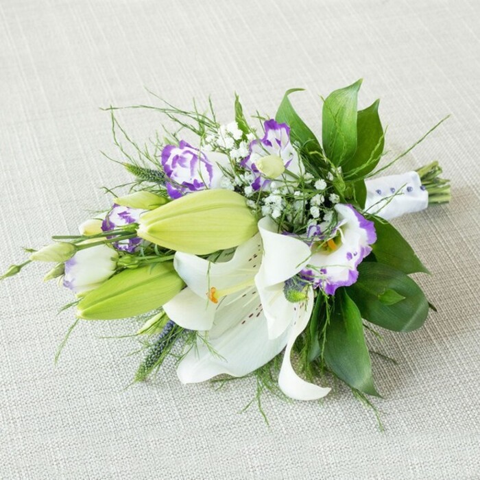 A graceful, hand-tied bouquet stretches diagonally across a subtly textured light beige fabric, as if it has just been set down on a linen-covered table in a Grove Park home. Dominating the arrangement are large white lilies, some fully open and some just beginning to unfurl. Their petals are smooth and slightly curved at the tips, revealing tall, yellow-orange stamens dusted with pollen and a faint constellation of reddish-brown speckles inside the throats, details that give each bloom its own personality. Nestled alongside them are numerous fresh lily buds, a lively chartreuse-green that contrasts beautifully with the creamy whites, suggesting this arrangement will continue to evolve over the coming days. Between the lilies, clusters of Lisianthus blooms weave through the bouquet. Their petals are layered and gently ruffled, creating cup-shaped flowers that shift from creamy white at the centre to bold purple or rich violet at the edges, reminiscent of the playful colour accents you might see in shopfronts along nearby high streets. Fine stems of baby's breath scatter tiny white blossoms throughout, creating a gauzy, cloud-like veil that softens the stronger lines of the lilies and Lisianthus. A generous array of foliage frames the entire piece: broad, glossy dark green leaves provide depth and form, while wispy, more delicate greenery and a few subtly spiky elements add movement and dimension, almost as if caught in a soft Thames-side breeze. At the base, the stems are pulled together and secured with a smooth white ribbon, embellished sparingly with pearl-like beads that glimmer where the light catches them, hinting at bridal parties, engagements, or elegant celebrations held in and around Grove Park. The lighting in the image is gentle and diffuse, without harsh shadows, allowing the natural textures and fresh colours to shine through with understated sophistication.