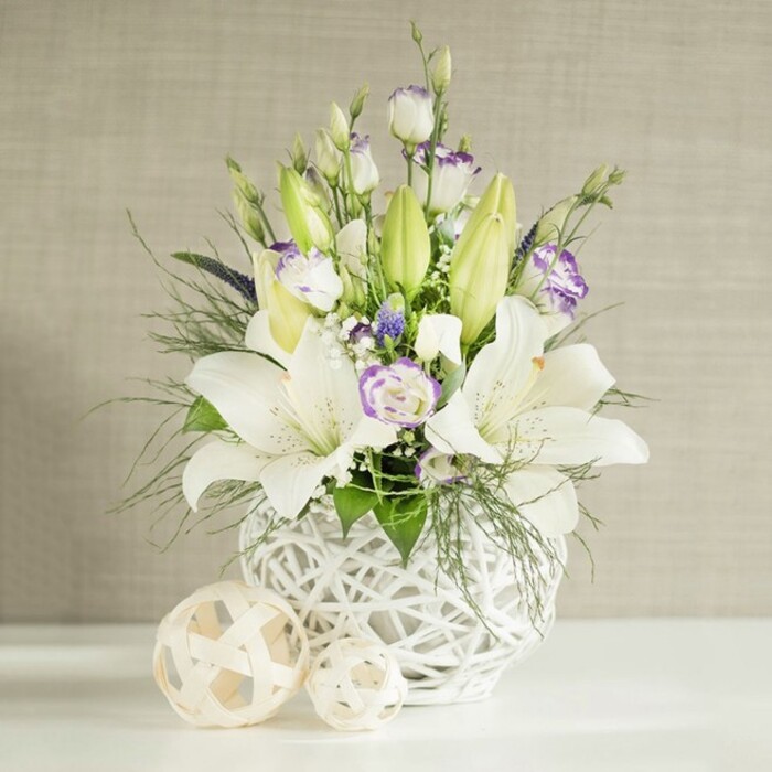A luminous bouquet of white lilies and lisianthus, expertly styled for a wedding table, stands in a sculptural white vase against a softly textured beige backdrop, evoking the gentle pace of life around Grove Park. The vase itself is round and slightly oversized, its open-weave structure resembling finely crafted wickerwork in ceramic form, creating airiness beneath the flowers. From this base, several graceful lilies rise; some blossoms are fully open, their long petals curving outwards to reveal central stamens dusted in yellow pollen, while others remain sequestered in smooth, green buds that add vertical shape and a sense of anticipation. Interspersed among the lilies are numerous lisianthus blooms, their petals layered and gently ruffled, predominantly white with subtle lavender-purple tinges along the edges or at their centres, adding a soft wash of colour. Fine sprays of small white flowers, similar to baby's breath, and a scattering of slightly deeper purple blossoms, possibly reminiscent of muscari, are nestled within the design, enhancing texture and creating depth. Surrounding the blooms, a combination of feathery foliage and broader green leaves extends and drapes over the rim of the vase, suggesting movement and giving the impression that the bouquet was freshly gathered from gardens around Chiswick and Grove Park. Resting on the clean white surface next to the vase are two woven decorative spheres in light beige and natural wood tones, echoing the vase's pattern and contributing to the cohesive, contemporary aesthetic. Behind, a light beige wall with a horizontal, linen-like texture is bathed in even daylight, softening any shadows and allowing the whites to appear crisp and pure. The scene conveys serenity, purity, and thoughtful elegance, making the arrangement perfect for wedding receptions, anniversary dinners, or meaningful sympathy tributes across Grove Park.