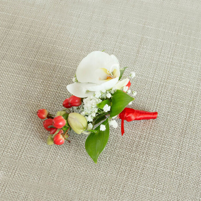 Resting on a pale, wheat-toned linen fabric, this refined floral boutonniere is captured in a gentle overhead view that reveals every careful detail a local Grove Park florist might consider before handing it over on a wedding morning. At the heart of the design is a singular white Phalaenopsis orchid, its petals wide and slightly cupped, with a soft, velvety texture that almost suggests cool porcelain to the touch. In the centre of the orchid, subtle tones of creamy yellow and soft orange glow within the throat, creating a warm focal point that draws attention without overpowering the composition. Circling this main bloom, tiny white blossoms of baby's breath form a delicate, airy frame, their many small florets lending dimension and a light, cloudlike quality. Along one side, a cluster of round, glossy red berries provides a bold splash of colour, reminiscent of winter hedgerows around nearby Chiswick and Strand-on-the-Green. Interspersed among them, a few lighter green, unripe-looking buds bring variety and a sense of evolving growth. Rich green foliage, in a mix of broader, pointed leaves and slimmer, elongated shapes, weaves through the flowers and berries, giving structure and a natural, garden-fresh feel. The stems are firmly wrapped at the base with a bright red satin ribbon, its smooth sheen catching the light as it forms a compact knot, with short ends that lie neatly along the fabric. The neutral, woven backdrop keeps attention on the interplay of white, red, and green, allowing the textures - from satin to petal to leaf - to stand out clearly. This small floral piece feels perfectly suited to a groom or groomsman at a Grove Park ceremony, a seasonal formal event, or a holiday gathering, offering a quietly luxurious accent that sits close to the heart.