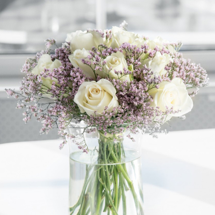 An elegant glass vase filled with white roses and lavender-toned filler flowers creates a serene sympathy display, captured in a close-up that invites you to linger over the details. The roses are fully open, their creamy white petals arranged in concentric spirals that slowly expand outward, forming soft, rounded blooms with slightly ruffled edges. They are spaced so that each flower can be appreciated, yet close enough to form a lush, continuous canopy of white. Nestled among these roses are clusters of fine, misty filler blossoms in shades of pale lavender, lilac, and the softest pink, most likely statice, their small, papery petals catching the light and adding a gentle, cloud-like texture. The bouquet rests in a tall, cylindrical clear glass vase, where the neat green stems are visible all the way down into clean, fresh water, a sign of the care taken by Grove Park Florist in preparing each stem. The vase is placed on a pristine white surface that reflects the underside of the arrangement, giving a sense of lightness and floating calm. The background is softly blurred, a subtle haze of white and grey tones that suggests a bright, peaceful interior-perhaps a living room or hallway in a Grove Park home, just a short walk from the quiet corners of local parks or the leafy streets leading toward Chiswick. Natural light washes gently over the bouquet, bringing out the creamy highlights in the roses and the muted purple of the filler flowers. Overall, the image conveys grace, simplicity, and quiet compassion, perfectly suited to moments of condolence and remembrance.