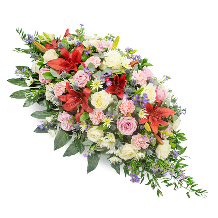 The image shows a substantial, artistically composed casket spray laid out diagonally against a stark white background, allowing every nuance of colour and detail to be clearly appreciated. The arrangement begins with a rounded mass of blooms at the broader end and narrows gently to a soft, pointed finish, creating a graceful line that suggests stillness and repose. Dominating the design at intervals are deep red lilies with large, star-shaped petals, acting as striking focal notes that convey depth of feeling and respect. Around and between these lilies, a blend of white, cream, and pale pink roses unfolds, some blossoms fully open with velvety, layered petals, others still cupped in bud, giving the impression of time passing and memories layered one upon another. Light pink carnations, their edges finely frilled, mingle with smaller white chrysanthemum-type flowers, building up a generous, cloud-like fullness across the centre. Scattered among them, blue-purple thistles and clusters of small lavender-blue filler blooms, likely veronica or statice, add cooler tones and tactile contrast, like soft echoes of the riverbank and parkland flora found near Grove Park and the Thames. A lush foundation of deep green foliage supports the flowers: broad, shiny leaves provide visual weight and structure, while more delicate, fern-like greenery trails and extends beyond the blooms, particularly at the narrower end, giving the piece a gentle sense of movement. The neutral white background and soft, even light ensure that shadows are minimal and the colours remain true. Overall, the spray feels like a tender, considered tribute a family might choose for a farewell service in the Grove Park area, designed to express enduring love, sympathy, and quiet reverence.