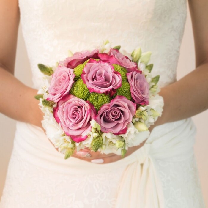 From the refined lace of the bride's dress to the vibrant layers of expertly chosen flowers, this close-up portrays a classic Grove Park bridal bouquet with a fresh, romantic twist. The bride's arms, gently holding the compact arrangement, are set against the delicate pattern of her ivory gown, which features lush lacework and a subtle, unadorned drape at her waist-a familiar sight in elegant Grove Park weddings. At the bouquet's center, several large roses bloom in shades of lavender and mauve, their petals deepening to fuchsia or magenta at the edges for dramatic contrast. These opulent roses are surrounded by a lively ring of lime-green button chrysanthemums, whose textured, pom-pom shapes bring playful energy reflective of Grove Park's vibrant green spaces. Completing the design is a crown of tiny white blossoms-possibly spray roses or freesia-interspersed with green buds, infusing the bouquet with gentle charm and a sense of lightness. The bride's polished nails, delicately refined with a pink shimmer, and the gentle, even lighting create an inviting, serene mood. Crafted by Grove Park Florist, this arrangement beautifully weaves together classic and modern floral elements, making it a cherished symbol of love and artistry for the Grove Park community and beyond.