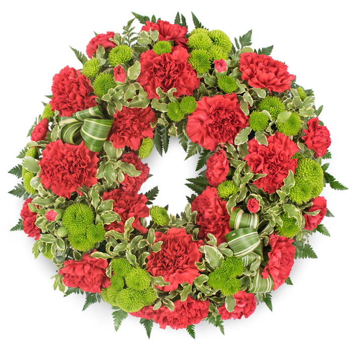 A lush circular funeral wreath is displayed front-facing on a stark white background, creating a vivid contrast that highlights every flower, leaf, and ribbon detail. The wreath itself forms a perfect ring, completely filled with a combination of deep red carnations and lime green button chrysanthemums, ensuring that the entire surface is richly textured and visually full. The carnations provide the foundation: generous, multi-layered blooms with frilled edges that catch the light and give each flower head a soft, almost fabric-like appearance. Their crimson colour runs consistently throughout, from larger open blossoms to smaller buds, establishing a sense of warmth and solemnity. Punctuating this red are the lime-toned button mums, their rounded, compact domes of tiny florets offering a smooth, modern counterpoint to the carnations' more intricate petals. Together, the two types of flowers create a striking play of texture and colour, red and chartreuse weaving in and out around the circle. Beneath and around them, a structured bed of greenery lends depth: dark green leatherleaf fern adds a feathery, layered backdrop, while variegated pittosporum with pale, creamy edges gently lifts the edge of the wreath, giving it a lighter, more dimensional outline. Interlaced with the foliage are graceful loops of striped green ribbon, echoing the fresh tones of the button chrysanthemums and adding a handcrafted, personal touch. The wreath feels thoughtfully designed, symbolising an unbroken circle of memory and love. It evokes the kind of respectful tribute a Grove Park florist might arrange for services at local churches or for delivery to families in Grove Park, offering a dignified, comforting expression of sympathy.