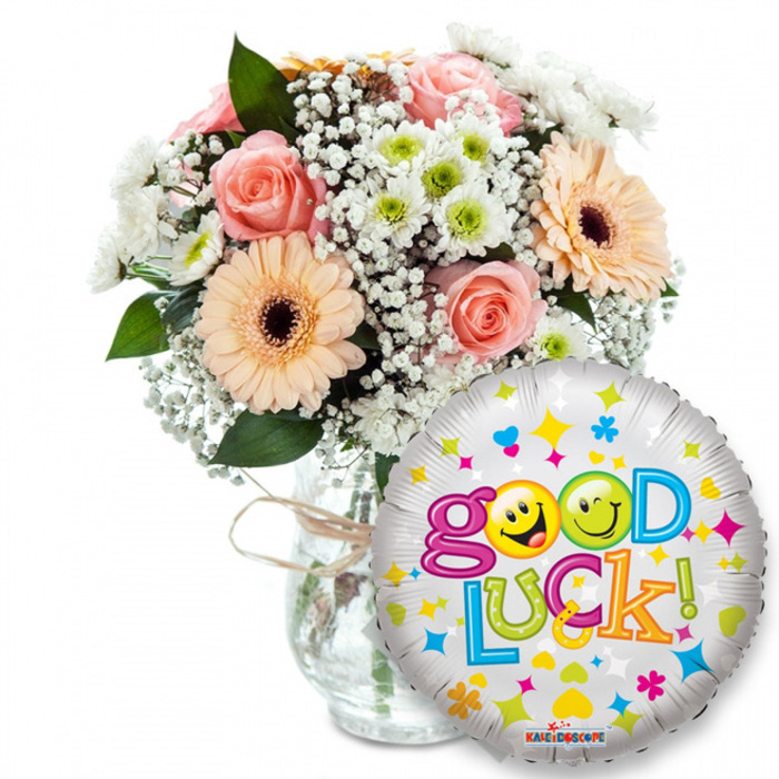 Framed by a simple white backdrop, this uplifting 'Good Luck' gift set feels intimate and hand-crafted: a lush bouquet stands in a classic clear glass vase, cinched with rustic twine, the knot hinting at an artisan finish. The blooms are a gentle study in soft tones - peach-orange Gerberas with dark brown centres provide cheerful, satiny petals, while light pink roses offer rounded, velvety depth. Wisps of white baby's breath spill through the composition, and small, crisp white-and-lime chrysanthemum florets add bright, textured accents. Deep green foliage pushes through the blossoms, creating shadowed pools that make the pastel tones sing. In front, a shiny round silver foil balloon overlaps the arrangement, its metallic face scattered with colorful confetti motifs and crowned by the jubilant phrase Good Luck! in bubbly, multicoloured type. The two o's in Good are replaced by yellow smiley faces - one winking, one tongue-out - lending a playful, human touch; the KALEIDOSCOPE logo sits near the balloon's base. The overall impression is quietly celebratory: you can almost sense the cool Mylar against fingertips and the faint rose scent in the air. This arrangement is suited to send from a local neighbourhood florist in Grove Park - perhaps delivered to a flat near Grove Park station or left on a windowsill overlooking the nature reserve - to mark exams, interviews, farewells or hopeful new beginnings.
