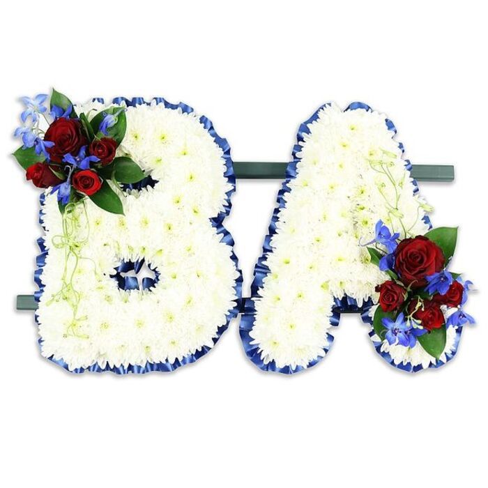 A carefully composed memorial piece forming the uppercase initials 'B' and 'A', each letter densely covered in hundreds of pristine white chrysanthemum heads to create a smooth, pillowy texture and an overall sense of serenity. The edges of both letters are delineated by a sumptuous royal blue satin ribbon that has been ruffled slightly to give subtle depth and a crisp silhouette against the white field. At the upper left of the 'B' an arresting floral cluster brings rich colour: deep scarlet roses in mixed stages of bloom nestle with lively, intensely blue delphinium-like blossoms, while soft, pale-green trailing foliage and darker green leaves provide counterpoint and movement. A matching cluster graces the lower right of the 'A', repeating the same velvety red roses, electric blue spikes, and lush greens so the pair read as one lovingly balanced tribute. The two initials are affixed side-by-side on a discreet dark green rectangular stand, giving stability and a low-profile base that visually anchors the piece on a chapel table or memorial plinth. The neutral white background in the photograph accentuates the saturated red and blue accents and the delicate layering of textures. This bespoke, lettered tribute is the kind of sympathy piece chosen for funerals and commemorations to personalise remembrance; I've placed similar designs for services around Grove Park and nearby Downham, where families appreciate the combination of quiet white chrysanthemum purity with bold, sentimental accents.