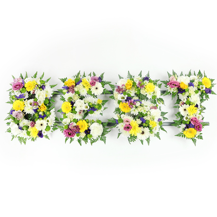 This photo presents a handcrafted floral tribute spelling out BEST in four bold letters, centred horizontally on a pristine white background and photographed head-on with even studio light that keeps every petal crisp and true to tone. The arrangement balances cool and warm notes: pristine white chrysanthemum-like blooms and cream gerberas offer a serene, pure base while yellow roses and gerbera daisies inject bright optimism. Interspersed throughout, lilac and deep-purple lisianthus or smaller chrysanthemum blooms bring soothing shadow and visual depth; vivid fuchsia-pink spray roses or carnations lift the mood with vibrant personality. Small green and white filler clusters add a dewy texture between the larger blooms, and broad, dark-green fern fronds weave around the letters' perimeters, creating a soft, organic halo that helps each letter read clearly from a distance. A subtle dark-green or grey metal framework supports the lower bars-noticeable in the E and the T-but never overwhelms; instead it gives the piece structural grace. The scent is implied rather than depicted: a quiet mix of rose sweetness and the fresh greenness of cut foliage. This composition is suited to memorial services, tributes, or occasions when a bold, worded statement is needed; locally, families in Grove Park, near Chinbrook Meadows and the station, often choose this style when they want a message that is both personal and unmistakable, conveying warmth, respect, and celebration of a life remembered.