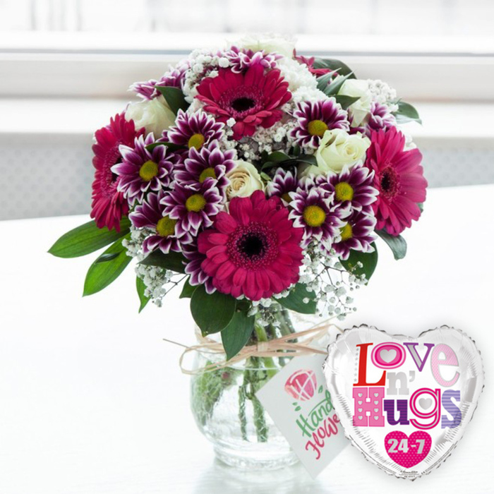 A joyful floral arrangement in a clear glass vase rests on a pale, uncluttered surface, set against a softly blurred, bright background that suggests a sunlit room in Grove Park. The bouquet has a full, rounded shape, built around several large gerbera daisies whose petals glow in rich magenta and fuchsia, radiating from deep, almost black centres that draw the eye. Interwoven among these statement flowers are numerous smaller chrysanthemums with a distinctive two-tone look: velvety purple on the inner part of each petal, lightening to a clean white at the edges, and finished with a bright yellow centre that gives each bloom a cheerful, almost playful expression. Creamy white roses sit harmoniously between these vivid colours, their gently curled petals and classic form introducing a softer, romantic note. Delicate sprays of white baby's breath float throughout the bouquet, softening transitions between the larger blooms and adding a fine, lacy texture that makes the arrangement feel light despite its abundance. Lush green foliage peeks through at different heights and angles, grounding the composition and evoking the greenery of nearby parks and gardens around Grove Park. The stems are visible through the clear vase, carefully cut and arranged in fresh water, showcasing the thoughtful craftsmanship of a skilled florist. Around the neck of the vase a band of rustic twine is tied, and from it hangs a small, white gift tag bearing a stylised pink rose motif and informal green lettering, hinting at a hand-finished, personalised gift. Just to the right, leaning comfortably against the vase, a heart-shaped foil balloon adds a festive touch. Its silver border gleams, while the white interior is decorated with colourful mosaic-style letters in pinks, purples, and blues spelling out the affectionate message Love n' Hugs 24-7, punctuated with tiny hearts. The whole scene, washed in bright, natural light, feels warm and inviting, perfect for expressing love, marking a special milestone, or brightening someone's day with a same-day delivery from Grove Park Florist.