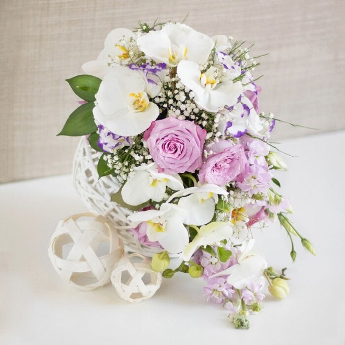 An exquisitely detailed cascading bouquet stretches diagonally across the frame, captured against a soft off-white backdrop that recalls a sunlit Grove Park reception room just off Burlington Lane. The arrangement is styled in a classic waterfall or teardrop silhouette, with the bulk of the blooms gathered near the top and tapering down into a graceful point. At its core, several pristine white orchids-similar to Phalaenopsis-anchor the design; their broad, slightly arching petals and gentle yellow centers add an air of polished sophistication. Surrounding these orchids, roses in delicate blush pink and muted lavender unfold at various stages of bloom, their petals layered and slightly ruffled, giving the bouquet a lush, romantic texture. Delicate stems of white baby's breath weave between all the flowers, filling spaces with a fine, cloud-like spray that softens the edges and imbues the composition with an ethereal lightness. Subtle strands of trailing blooms and greenery extend downward, enhancing the sense of flowing movement and guiding the eye along the curve of the cascade. Sprigs of fresh green foliage, carefully tucked among the blossoms, give dimension and a natural garden feel without interrupting the soft colour harmony. The bouquet rests upon or emerges from an intricate white decorative support-its surface appearing lace-like or embossed-and around it lie several small white or cream spheres, each with ornate cut-out patterns that echo the delicacy of the flowers. These sculptural accents help frame the bouquet and suggest a carefully curated wedding table setting. The background remains gently blurred and warmly lit, allowing the pastel roses, pristine orchids, and airy baby's breath to stand out clearly, a perfect visual for couples planning elegant celebrations in and around Grove Park. Crafted with the sensitivity of a local florist, the scene captures both opulence and quiet intimacy.