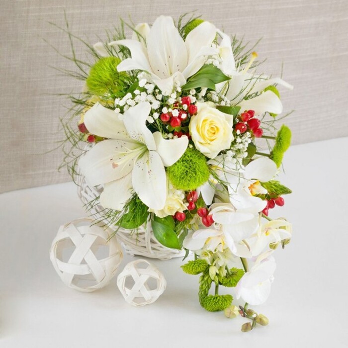 A richly detailed floral arrangement sits proudly on a smooth white surface, the clean lines of the table contrasting with a warm, textured beige background reminiscent of soft linen. The flowers are nestled into a white woven wicker basket, its round shape and intricate pattern anchoring the display. Rising from this base, several large white lilies take center stage, their trumpet-shaped blooms fully open, revealing gracefully curved petals with subtle freckling and elongated stamens dusted with pale golden pollen. Just above the middle of the bouquet, a single cream to pale yellow rose opens in a perfect spiral, its velvety petals drawing the gaze like a quiet point of light. From the right side, an elegant cascade of white phalaenopsis orchids flows downward, each stem bearing broad, smooth petals and a refined central column, creating a waterfall effect that would look stunning on a wedding table in a Grove Park venue near the Thames. Around these focal flowers, bright green pom-pom chrysanthemums cluster in rounded tufts, adding an almost moss-like texture and lively contrast. Interspersed among them are fine, wispy leaves and feathery greens, together with slightly broader, glossy foliage that adds shape and volume. Tiny white baby's breath blooms float throughout, giving the whole piece an airy, ethereal quality. Small clusters of vivid red berries punctuate the design, providing touches of rich colour that feel festive yet balanced. To the left on the white tabletop, two decorative spherical ornaments made from white woven material-one larger, one smaller-sit alongside the basket, their open, geometric latticework echoing the basket's weave and adding sculptural interest. The overall mood is calm, luxurious, and celebratory, an expertly crafted arrangement of fresh flowers that could serve as a wedding, anniversary, or special occasion centerpiece for homes and venues around Grove Park.