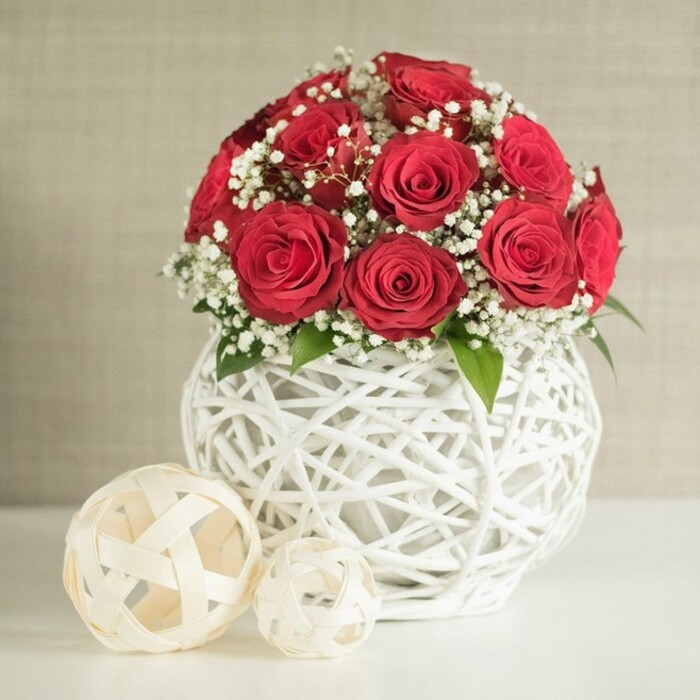 A beautifully composed floral still life captures the essence of a romantic wedding table arrangement, as though set for an intimate reception in Grove Park. At the centre is a lush, rounded bouquet of deep crimson roses, numbering around a dozen to fifteen. Each rose is in full bloom, with tightly layered petals that spiral inward, giving the impression of velvety, three-dimensional rosettes. Interspersed among these rich red flowers are gentle clusters of white baby's breath, fine stems branching into tiny blossoms that sit like small, weightless clouds against the darker petals, lending the arrangement softness and light. Peeking subtly from beneath and between the blooms are a few green leaves, minimal yet crucial, adding hints of freshness and helping define the natural contours of the bouquet. The flowers are gathered into a distinctive white vase with a rounded, bulbous shape and a highly textured open-weave design, crafted from slim, white interlaced strands. This lattice structure, reminiscent of handwoven wicker given a modern twist, creates depth, shadow, and a sense of airy lightness beneath the dense crown of roses. The vase stands on a pristine white surface, likely a tabletop or dressed wedding table, its simplicity ensuring that the arrangement remains the focal point. Just to the left of the vase are two decorative woven spheres, fashioned from flat, cream-coloured strips that form open, basket-like orbs; the larger sphere is positioned closer to the viewer, while the smaller sits partly behind, their shapes and textures harmonising with the vase. In the background, a softly textured wall in pale beige fades into gentle blur, providing a calm, neutral backdrop that accentuates the vivid reds and crisp whites. The overall effect is one of poised elegance and romantic simplicity, perfectly suited to describing wedding centrepieces, anniversary dinners, or refined home decor in the Grove Park area.