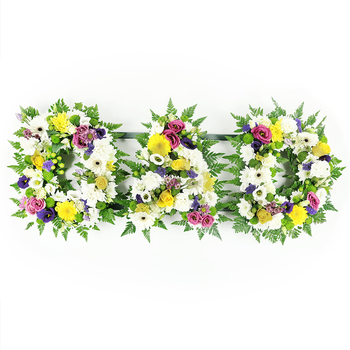 This photograph features a classic DAD funeral tribute presented as three large floral letters, laid out in a neat row against a plain, bright white backdrop. Each letter is fashioned on a generous bed of deep green fern foliage, the fronds radiating softly around the shape and adding gentle movement to the outline of the first D, the A in the centre, and the second D on the right. The faces of the letters are completely covered in white blossoms, primarily chrysanthemums and wide-headed gerbera daisies, arranged so closely that they form a plush, velvety surface without gaps. The white flowers convey purity, peace, and remembrance, evoking the quiet atmosphere of a chapel or small family service in Grove Park. Woven through the white base are scattered clusters of yellow - sunny gerberas and small spray roses - which bring a sense of warmth and a nod to treasured memories and shared laughter. Rich purple lisianthus flowers and rounded magenta or deep pink spray roses sit among these, their stronger tones suggesting honour, affection, and respect. Small lavender flowers and pops of bright green button chrysanthemums add extra layers of texture and colour, like fine stitching in a well-crafted garment. At the back, slim dark green metal or plastic bars link each letter, ensuring the tribute stays straight and aligned when displayed on stands or beside a coffin. This DAD arrangement feels like something a family in Grove Park might order from their trusted local florist, wanting a floral tribute that is both visually striking and emotionally sincere, capable of expressing love, loss, and gratitude in a simple, dignified word.