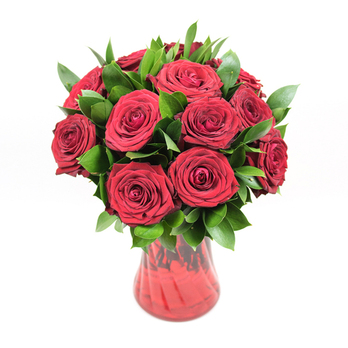 In this image, a sumptuous arrangement of around twelve to fifteen deep red roses is presented in a translucent red glass vase, set cleanly against a stark white background that makes every petal and leaf appear almost sculptural. The roses are the star of the composition: each bloom is fully open, with countless velvety petals layering tightly towards the centre and softening into larger, gently curved edges on the outside. The colour is a rich, classic crimson that feels both dramatic and tender, evoking the kind of enduring romance people in Grove Park might choose to celebrate a milestone anniversary or an important apology. Nestled among the blooms is a full collar of fresh dark green foliage, likely ruscus or salal, with smooth, slightly oval leaves that peek between the roses and extend just beyond the edge of the arrangement. This greenery adds body and shape, creating a rounded silhouette and preventing the bouquet from feeling too formal, instead giving it a natural, garden-inspired charm. The vase itself is a deep, matching red tone, with a flared opening and a softly curved body that narrows elegantly towards the base. A faint ribbed or swirling texture runs through the glass, catching the studio lighting so that fine highlights and shadows ripple across its surface, echoing the folds of the petals above. The stems of the roses are only subtly visible through the glass, hinting at the careful, spiralled placement that a local florist, such as Grove Park Florist on the high street, would use to keep the bouquet perfectly balanced. The clean, uncluttered background and even, bright lighting create a sense of calm and focus, making this bouquet feel like a still moment of affection and devotion, ready to be carried through the familiar streets of Grove Park to someone dearly loved.