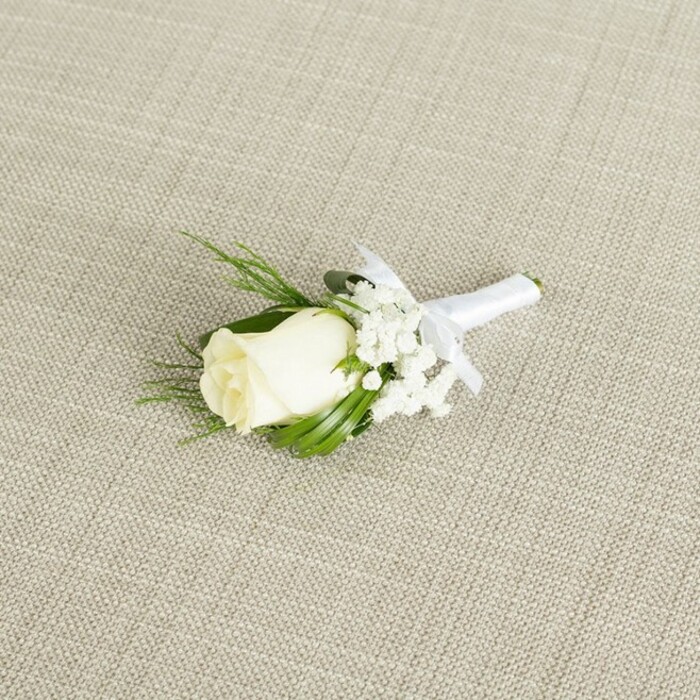 An elegant white rose boutonniere lies at a diagonal angle on a neutral, light beige fabric that looks like finely woven linen, its subtle texture providing an understated backdrop reminiscent of calm interiors in Grove Park homes. At the centre of the composition is a single ivory rosebud, medium in size, its petals still closely cupped yet already showing the gentle curves and layers that will unfurl with time. The rose has a soft, creamy white tone with a hint of warmth, creating a sense of purity without feeling stark. Just beneath the bloom, touches of pale green from the sepals and stem suggest freshness, as if the flower has only recently been prepared. Encircling the rose are delicate tufts of white baby's breath, their numerous tiny blossoms resembling miniature stars or clouds, bringing a light, romantic airiness that softens the whole piece. Several narrow, pointed green leaves and a few more textured, feathery sprigs of foliage extend around the central flower, framing it like a natural wreath and emphasizing the contrast between deep green and luminous white. The stems have been carefully gathered and wrapped in smooth white satin ribbon, the wrap perfectly tight and even, culminating in a small, symmetrical bow positioned neatly just under the flower heads, signalling that the piece is ready for a groom or groomsman's lapel. Gentle, diffused lighting falls across the scene, enhancing the satiny sheen of the ribbon, the plush quality of the rose petals, and the airy intricacy of the baby's breath. Crafted with the attention to detail you might expect from an experienced local florist serving weddings throughout Grove Park and the nearby riverside, this boutonniere radiates quiet elegance, tradition, and the tender excitement of a wedding day about to begin.