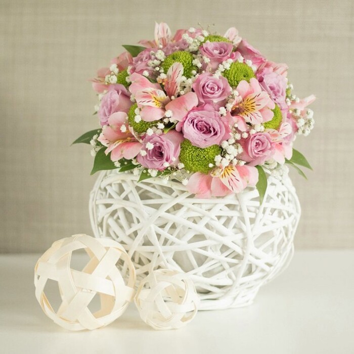 A voluminous floral arrangement stands proudly on a spotless white surface, its rounded silhouette framed by a warm, textured beige backdrop that feels like the painted walls of a calm Grove Park sitting room. The centerpiece is cradled in an eye-catching white vase, crafted from slender strips woven into an open, basket-like lattice. The vase swells gently outward toward the middle before narrowing at the top, giving it a sturdy, comforting presence, as if it has been placed deliberately in the heart of the home. Rising from this sculptural base is a lavish cluster of roses in pastel pink and soft lavender, the blooms fully opened to reveal layers of velvety petals that curl and fold in on themselves, catching the soft studio light in subtle highlights and shadows. Nestled between the roses are graceful stems of pink alstroemeria, their trumpet-shaped flowers opening wide, each petal streaked with fine, darker lines and hints of creamy yellow deep in the throat, adding a delicate sense of movement and patterned detail. Breaking up the romance of the pinks and purples are playful lime green button chrysanthemums, small, spherical, and densely petalled, dotting the bouquet with vivid energy that feels fresh and modern-perfect for a bright Grove Park breakfast nook or a stylish home office. Interlaced among all these blooms, sprigs of white gypsophila float like mist, their tiny flowers creating a soft halo that makes the composition seem almost weightless. Darker green leaves and foliage peek out from the base and through the middle, offering depth, contrast, and the impression of freshly gathered garden stems. To the left of the vase, two decorative spheres made from light, natural-coloured woven material rest on the table, one slightly larger and set closer to the flowers, the smaller edged forward. Their open, airy weave echoes the structure of the vase and introduces an element of thoughtful interior styling. The whole scene feels serene, refined, and welcoming, the kind of floral design Grove Park Florist might prepare for an intimate dinner, a soft spring celebration, or a gentle housewarming gift delivered to a neighbour just off Grove Park Road.