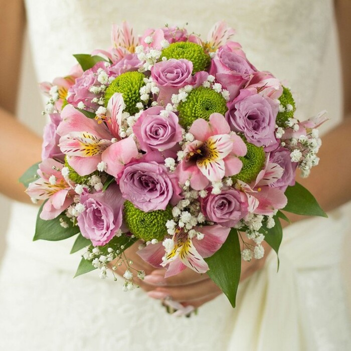 In the heart of Grove Park, where gentle breezes carry stories down Chiswick's leafy lanes, this bridal bouquet encapsulates the essence of local romance and expert floristry. Held lovingly by a bride in a luminous white gown, the bouquet's rounded form glows with soft pink and lavender roses, their petals layered like poetry and glistening with a hint of dew. Adding lively energy, spherical button chrysanthemums in fresh lime green punctuate the design, mirroring the vibrant flora dotting Grove Park's open spaces. Alstroemeria lilies unfold between the roses, their trumpet-shaped petals painted in blush pink, streaked with maroon, and touched with sunny yellow - each bloom recalling quiet walks through Duke's Meadows. Wisps of white baby's breath encircle the arrangement, casting a cloud-like veil that softens the exuberance of the main flowers, while glossy dark green leaves offer foundation and contrast. The bride's hands cradle the bouquet securely, a simple band on her finger capturing the sweetness of commitment felt at local ceremonies. Every detail, from the intricate lace backdrop to the harmonious layering of blooms, reveals the artisan touch of Grove Park Florist - a bouquet crafted with the spirit and vibrancy of the community in mind. Designed for spring or summer weddings and memorable Grove Park events, this creation celebrates love, artistry, and the fresh promise of each new beginning.