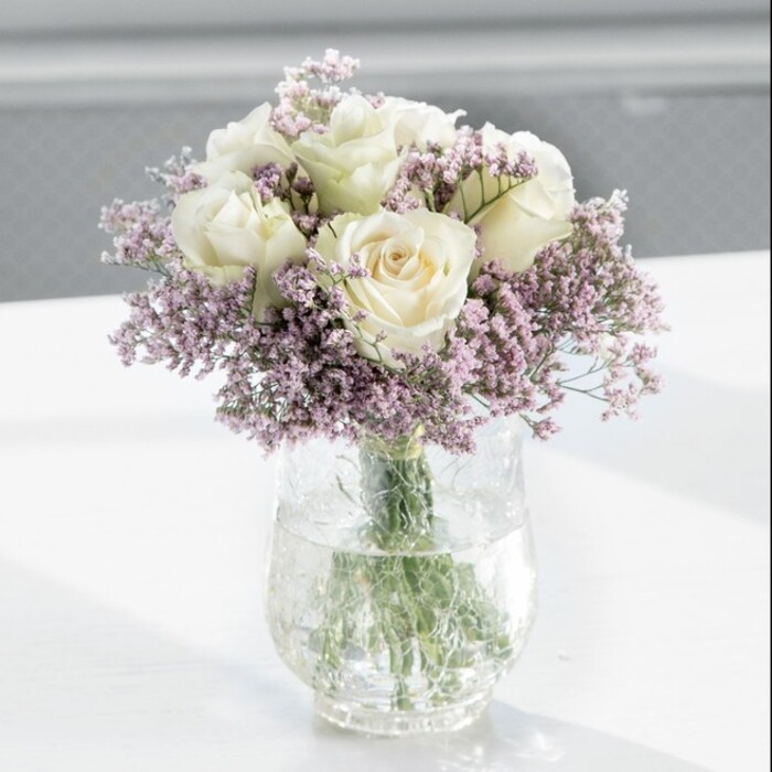 An elegant bouquet of creamy white roses and delicate lilac statice stands tall in a clear glass cylinder vase, the composition poised and balanced as if just styled for a Grove Park townhouse or a riverside flat near Chiswick and the Thames. At its core, around twelve white roses form a generous, domed cluster. Their petals, thick and velvety to the eye, curl softly outward in perfectly formed spirals, the colour hovering between soft cream and polished ivory. Each bloom feels intentional, placed to create a smooth, rounded silhouette with no harsh gaps or sharp angles. Threaded throughout, feathery sprays of light lavender statice weave in and around the rose heads. Each fine stem is crowned with dozens of minute blossoms, giving the arrangement a frothy, almost cloud-like frame. The statice adds a gentle dusting of colour-a cool, dusty lilac that enhances the roses' warm cream tone without ever overpowering it. Together they create a look that is both classic and quietly modern, the kind of refined simplicity Grove Park Florist is known for. All the stems are neatly aligned in a tall, clear cylindrical vase, immersed in sparkling water that refracts the stems into soft, green lines. The vase rests on a pristine white surface, perhaps a kitchen island or office reception desk, where the natural light from a large window sweeps across the scene. That light gives the petals a soft sheen and paints the faintest shadows beneath the vase. The background is blurred and pale, suggesting an uncluttered room and allowing the flowers to command full attention. The mood is peaceful yet affectionate, ideal as a housewarming gift for a new flat near Grove Park station, a comforting sympathy arrangement, or a timeless centrepiece for a small, intimate celebration.