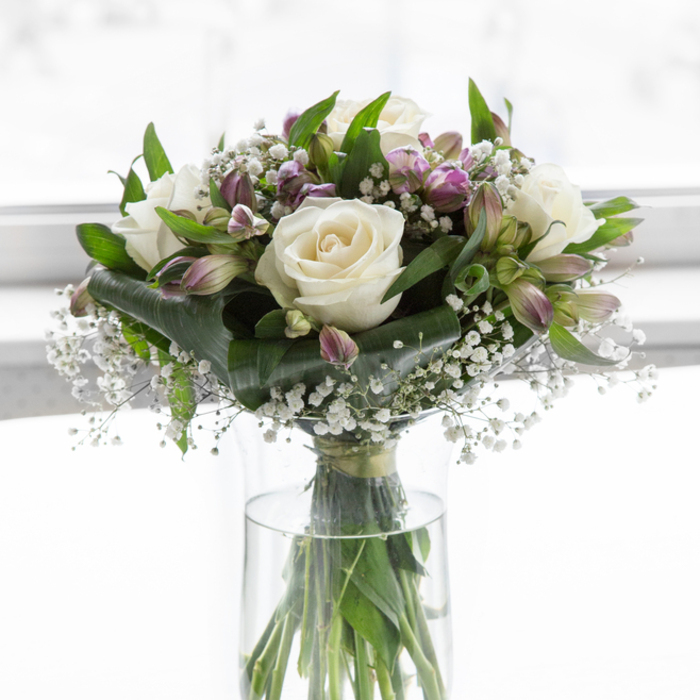 A softly luminous sympathy bouquet stands upright in a clear glass cylindrical vase, its gentle colours and textures highlighted by bright yet diffused light that feels like it could be streaming through a Grove Park bay window on a calm afternoon. At the centre, creamy white roses are arranged with care, some fully open to reveal their intricately layered petals, others just beginning to unfurl, giving a natural, unforced rhythm to the design. Their pure white tones convey a message of respect, love, and serenity. Intertwined with the roses are alstroemeria blossoms and buds, their slim petals washed in shades of lilac, soft lavender, and muted purple, with faint touches of green near the base, creating a subtle play of colour rather than a bold contrast. Delicate stems of baby's breath run throughout the bouquet, each one dotted with clusters of tiny white flowers that fill spaces softly, like a light mist drifting across the arrangement, adding an airy, almost weightless feeling. Around the blooms, a carefully chosen selection of greenery-broad, glossy leaves alongside finer, more elongated foliage-cradles the flowers and frames them, giving both structure and a sense of natural growth, much like the calm paths and green spaces around Grove Park station. Through the transparent sides of the vase, you can see the straight green stems neatly gathered in clear water, a quiet indication of freshness and attentive floristry. The backdrop is a bright, softly blurred white, devoid of any detail, which keeps the viewer's focus firmly on the bouquet itself. Altogether, this arrangement offers a refined, comforting presence, ideal for bringing a gentle note of solace to memorial services, condolence visits, or intimate spaces of reflection in the Grove Park community.