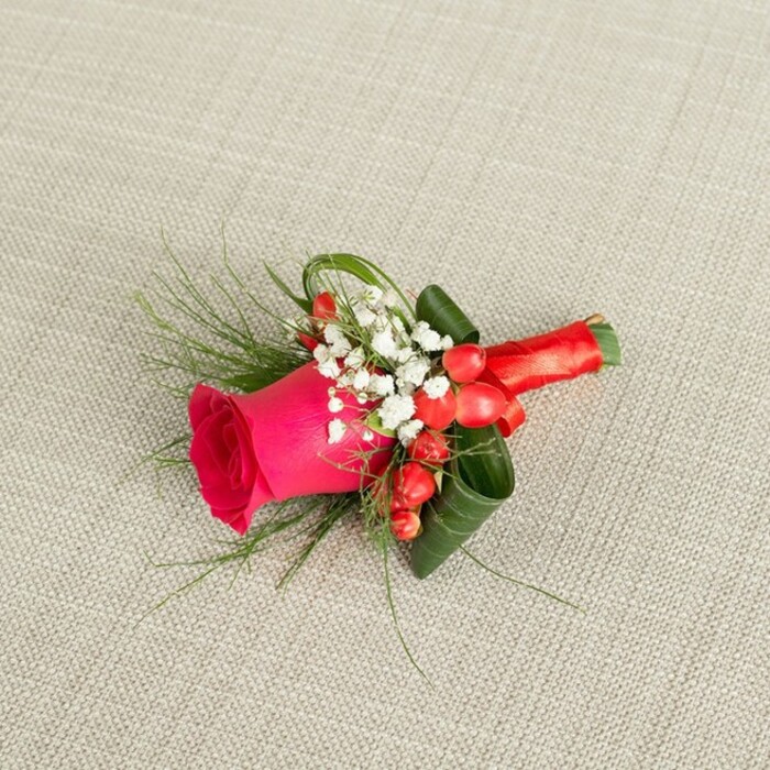 On a subtly textured, light beige woven fabric, this meticulously crafted boutonniere is positioned at a gentle diagonal, as if it has just been placed down after being pinned to a suit in a Grove Park bridal suite. The central star of the design is a single fuchsia pink rose, its petals densely layered and luxuriously velvety, opening just enough to show its fullness while still holding a sense of promise. Framing this rose are airy tufts of white baby's breath, each minuscule blossom creating a soft, mist-like border that lightens the overall look and provides a timeless, romantic counterpoint to the bold pink hue. Clusters of vibrant red hypericum berries nestle close to the rose, their round, polished surfaces catching the light, adding dimension and a festive pop of colour that hints at celebrations, toasts, and photographs taken along the quiet streets and green spaces of Grove Park. Fine, feathery sprigs of fern-like greenery extend delicately from the edges, while broader, darker green leaves are artfully curled and layered to give depth and structure, making the small piece feel both natural and intentionally designed. The stems are drawn together and tightly wrapped in a smooth, red satin ribbon, the finish precise and secure, suggesting the steady hands of an experienced florist. Gentle, diffused lighting casts soft shadows that emphasise the textures of fabric, foliage, and petals. The piece is clearly meant for a special occasion-a groom, groomsman, or prom date-offering a refined, romantic accent that would look perfectly at home at a Grove Park ceremony or reception, whether in a local hall, church, or a private garden celebration.