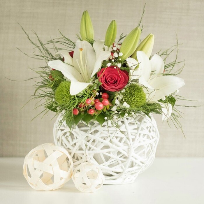 Set on a smooth white surface as if at the centre of an intimate wedding reception in Grove Park, this floral arrangement draws attention through its calm, balanced palette of white, red, and green. The flowers are gathered in a striking round white vase with a textural, open-weave construction, the interlaced strands forming an airy, basket-like sphere that feels modern yet organic. Rising from the vase, graceful white lilies create height and movement; some blossoms are fully open, their wide, slightly arched petals revealing small speckles and prominent golden stamens, while other stems are topped with sleek, unopened buds, promising ongoing beauty and fragrance throughout the celebration. Among the lilies, velvety red roses add depth and romance, with one particular rose nestled near the centre, its tightly folded petals forming a classic spiral that immediately draws the eye. Bright green, spherical blooms-resembling pom-pom chrysanthemums or dianthus-sit low in the design, adding soft, tufted texture and a fresh burst of colour evocative of the verdant pathways around Grove Park. Light sprays of white gypsophila are interwoven delicately among the larger flowers, their tiny blossoms giving a cloud-like softness. Scattered red berries gleam against the foliage, suggesting winter gatherings, Christmas weddings, or festive anniversary dinners. The greenery, a mix of fine, wispy fronds and broader, darker leaves, spills casually yet thoughtfully from the vase, contributing to an impression of natural abundance rather than strict formality. To the left, two woven decorative spheres in pale, natural hues rest on the surface, echoing the vase's lattice and framing the composition. Behind, a gently textured background in soft beige and light grey tones keeps the focus firmly on the flowers, allowing their colours, textures, and forms to create a serene yet celebratory mood.
