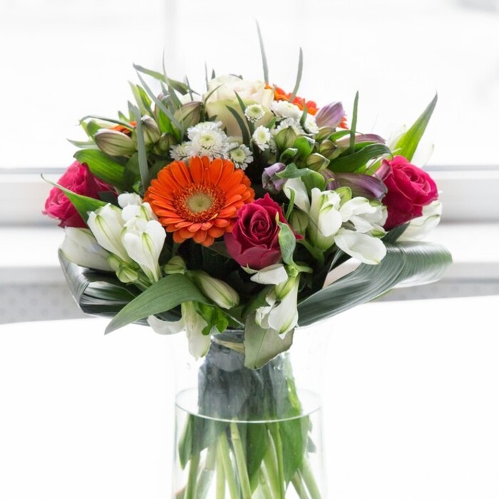 This exquisite bouquet is a demonstration of Grove Park's floristry expertise, full of visual poetry and soft vitality. Creamy peach gerberas radiate outward, encircling deep, fiery orange centres that bring to mind the vivid sunsets over Dukes Meadows. Interwoven with these are sumptuous fuchsia roses, their lush spirals imparting richness and gentle drama to the arrangement. Accents of ivory white alstroemeria and diminutive spray roses add a refreshing clarity, layered alongside delicate blush pinks and whisps of white filler flowers for extra dimension. The ensemble is embraced by wide, curving aspidistra leaves, their vivid green shine echoing the natural abundance found throughout Grove Park's neighbourhood parks. Smaller pointed leaves and fresh green foliage are artfully placed to harmonize the composition while adding hints of texture. Displayed in a slender, clear glass vase, every stem is visible-alive with colour beneath a wash of softly diffused daylight, much like morning light filtering through a local conservatory. Evoking togetherness, optimism, and new beginnings, this bouquet is equally suited for joyful gatherings, heartfelt congratulations, or simply expressing care for loved ones in Grove Park.