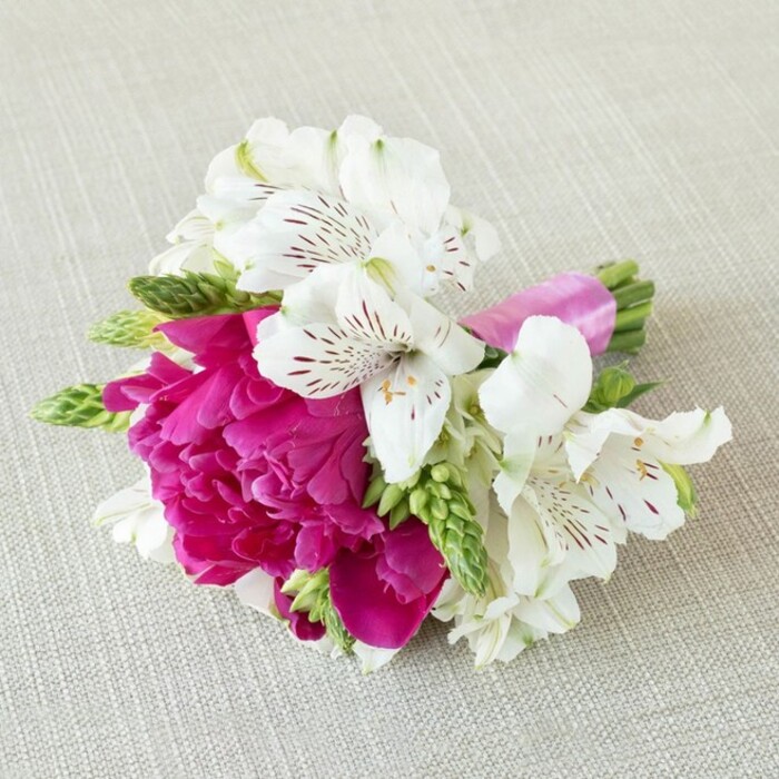 A beautifully detailed, compact floral piece rests on a softly textured, light beige fabric that resembles finely woven linen, the neutral background allowing every petal and ribbon to stand out with clarity. At the centre of this small bouquet is a single, richly coloured fuchsia bloom-peony-like in its fullness-with layers upon layers of ruffled petals spiralling inward, creating a plush, almost cushiony focal point. Cradling this vivid heart are several white alstroemeria blossoms, their trumpet-shaped petals slender and slightly curled at the tips, each one adorned with fine maroon streaks and speckles that radiate from the flower's throat like delicate ink lines. These white Peruvian lilies introduce an airy elegance and soften the drama of the bright pink. Threaded in between the larger flowers are slender stems of fresh green buds and foliage, possibly gladiolus or similar filler, their spiky, upright form and vibrant green hue adding structure, movement, and a refreshing, garden-like feel that evokes strolling through the leafy streets of Grove Park. The stems of the arrangement have been carefully aligned and tightly bound with a narrow band of glossy bright fuchsia satin ribbon, its smooth, reflective surface echoing the central bloom's vibrant tone and giving the design a polished, occasion-ready finish. Soft, even lighting falls across the scene, casting gentle shadows that accentuate the texture of the fabric backdrop and the subtle undulations of each petal. The overall impression is one of modern romantic charm, ideal as a boutonnière, corsage, or petite posy for an intimate wedding, anniversary dinner, or special celebration in one of Grove Park's cosy local venues, lovingly crafted with the eye of a florist who understands both colour harmony and emotion.