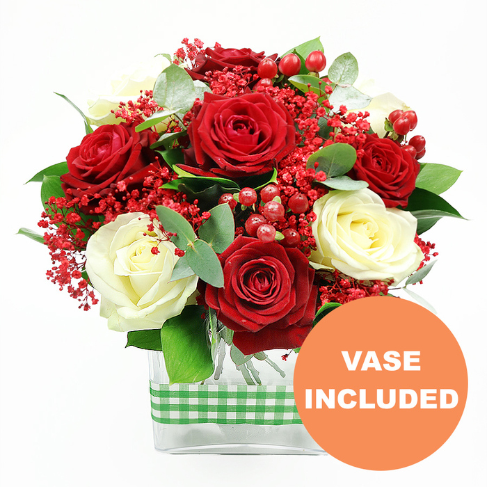 This image shows a sophisticated red and white rose arrangement set crisply against a clean white background, reminiscent of a carefully styled mantelpiece in a Grove Park townhouse. The bouquet is dominated by velvety deep red roses, each one open and full, their petals layered in soft, rounded spirals that suggest luxury and romance. Balanced against them are creamy white roses, their pale, smooth petals forming calm, rounded blooms that lighten the overall feel and introduce a sense of purity. Between these main flowers, the arrangement is laced with clusters of tiny, intense red filler blossoms, similar to tinted baby's breath, which create a haze of fine texture and help transition between the larger blooms. Dotted throughout are glossy red hypericum berries, each small sphere shining softly and adding a playful, decorative element that would be at home at a festive table or as a romantic gift delivered along Burlington Lane or near the Thames in Grove Park. Surrounding and supporting the flowers is an abundance of fresh green foliage, including slender, slightly curved eucalyptus stems and broader leaves that bring structure, depth, and an invigorating green contrast to the red and white tones. All stems are neatly contained within a clear rectangular glass vase, minimalist in design, allowing the natural pattern of the stems to show through and keeping the focus on the blossoms above. Around the centre of the vase, a green and white gingham ribbon is wrapped in a simple band, giving the piece a homely, rustic edge that recalls relaxed family gatherings. In the lower right corner, an orange circular emblem stands out against the white ground, with bold white text reading VASE INCLUDED, clearly indicating that the arrangement arrives complete and ready for instant display or same-day delivery from Grove Park Florist.