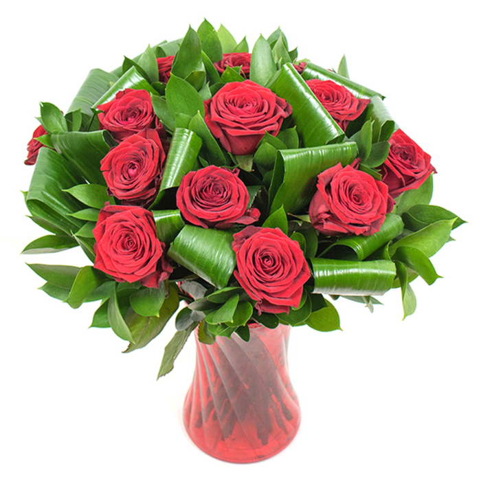 This portrait shows an elegant, densely packed bouquet of more than a dozen deep crimson roses, each in peak bloom with velvety, tightly coiled petals that gradually unfurl into rich, layered cups. The flowers possess a satiny sheen under soft, even lighting that reveals delicate ruffles and the tactile softness of the petals. Surrounding the roses is an abundant composition of dark green foliage: smooth, flat leaves provide a lush backdrop while broader glossy leaves are skillfully folded and rolled into dramatic spirals and arcs, introducing sculptural form and modern contrast to the otherwise classic rose display. The stems are gathered within a tall, slender, translucent red glass vase that carries a subtle twisted or ribbed texture; the vase's vertical lines catch and reflect light, reinforcing the bouquet's upright, architectural energy and amplifying the deep reds of the blooms. Placed against a crisp white background, the bouquet reads as a focused study in colour and texture, the reds and greens popping with clarity and presence. I can almost sense the delicate fragrance of garden roses mingling with the cool scent of fresh foliage - a composition that feels both passionately romantic and quietly sophisticated. This arrangement fits a variety of occasions: a refined anniversary gift, a Valentine's gesture, or a centerpiece for an intimate celebration near local landmarks like Grove Park green, Lee Road cafés, or the calm lanes toward Blackheath. The overall effect is timeless, luxurious, and thoughtfully composed, reflecting the careful hand of a neighbourhood florist who balances emotion with precision.