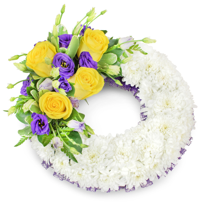 A meticulously arranged circular wreath occupies the frame, its principal form sculpted from a dense blanket of small white spray chrysanthemums that read as a seamless, plush ring. The massed white blooms, closely set and softly lit, create a tactile surface - powdered, yet resilient - that communicates purity and repose. From the upper left side a striking floral spray rises: luminous yellow roses in full bloom, petals unfurling from pale butter at the centre to richer gold at their edges, offer warmth and a faint floral note; they are balanced by clusters of deep purple lisianthus (Eustoma), whose satiny, ruffled petals lend a noble coolness and refined texture. A few unopened lisianthus buds in paler lavender introduce softness and a sense of unfolding life. Interspersed throughout are fresh green leaves - some glossy, some gently variegated - and fine fern fronds that separate and lift the colours, adding air and depth. Along the inner and outer curves of the white chrysanthemum ring a slim, dark-purple mesh trim peeks through, a subtle finishing touch that echoes the purple blooms and frames the composition. The wreath's open centre and classical circular form make it a traditional symbol of remembrance and condolence, intended for use at funerals or memorial services. This arrangement combines visual restraint with a comforting colour contrast, embodying a quiet farewell crafted with the measured care of an experienced neighbourhood florist near Grove Park station and local cemeteries.