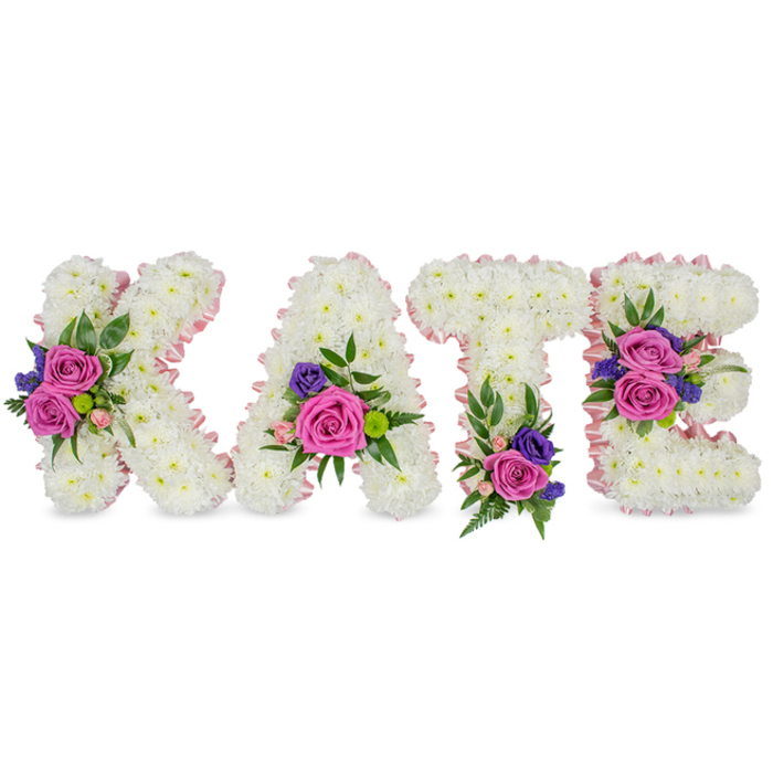 A carefully composed memorial spells KATE in four separate, upright letters, each built from a dense, uniform covering of immaculate white chrysanthemums that create a velvety, textural plane. A soft pale-pink satin ribbon delicately trims the outer and inner edges of every letter, introducing a subtle blush that frames the composition and gives each character gentle definition. Strategic clusters of colour punctuate the white: large fuchsia-pink roses bring warmth and affection, accompanied by smaller pastel pink spray roses for tenderness, and small deep purple or blue accents-statice or lisianthus-offer solemn contrast. On the K a prominent floral group sits on the diagonal stroke, the A carries blooms near its lower right curve, the T shows a cluster near its base, and the E bears two bouquets on the middle and bottom bars, providing a balanced rhythm when the tribute stands at a chapel or graveside. Varied greens-feathery fern fronds, smooth eucalyptus leaves and glossy foliage-frame each cluster, adding sculptural depth and a fresh, herbal scent that complements the rose sweetness. The overall palette of crisp white, soft pink, vivid fuchsia and deep purple reads as dignified yet personal under soft daylight or indoor ceremony lighting, projecting comfort and remembrance rather than spectacle. This style of name tribute is a classical choice for funerals, memorial services or celebrations of life-a bespoke expression of sympathy and gratitude, often chosen by local families across Grove Park, Lee and Downham who seek a sensitive, handcrafted floral memorial.