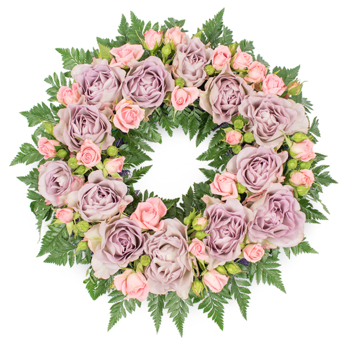 A meticulously arranged sympathy wreath lies flat on a spotless white surface, viewed from directly above so that every part of its circular design can be appreciated. The wreath is lavishly filled with roses, the largest of which are fully open blooms in muted tones of lavender, dusty purple, and soft mauve. These roses are richly petalled, their outer petals gently curving back while the inner whorls form intricate spirals, lending the arrangement a sense of depth and quiet luxury. Nestled closely between these prominent flowers are clusters of smaller, blush pink spray roses and delicate rosebuds. Some buds appear tight and compact, while others show the first signs of opening, their lighter pink tones adding a subtle touch of warmth and innocence to the more sophisticated lavender base. This interplay of size and stage of bloom creates a textured surface that invites the eye to move slowly around the circle. Beneath and beyond the flowers, a dense foundation of dark green fern fronds spreads outward, forming a textured frame. The ferns' fine, jagged leaflets peek between roses, adding dimension and a fresh, woodland quality that balances the formal elegance of the blooms. The lighting is bright yet soft, ensuring that the nuanced colours-from cool greyish purples to warm blush pinks-are clearly visible without glare. This wreath feels like a sensitive, carefully considered tribute, suitable for a funeral, wake, or celebration of life in Grove Park, where families might seek a floral expression of sympathy that speaks of serenity, gratitude, and cherished memories shared along the Thames paths and tree-lined avenues nearby.