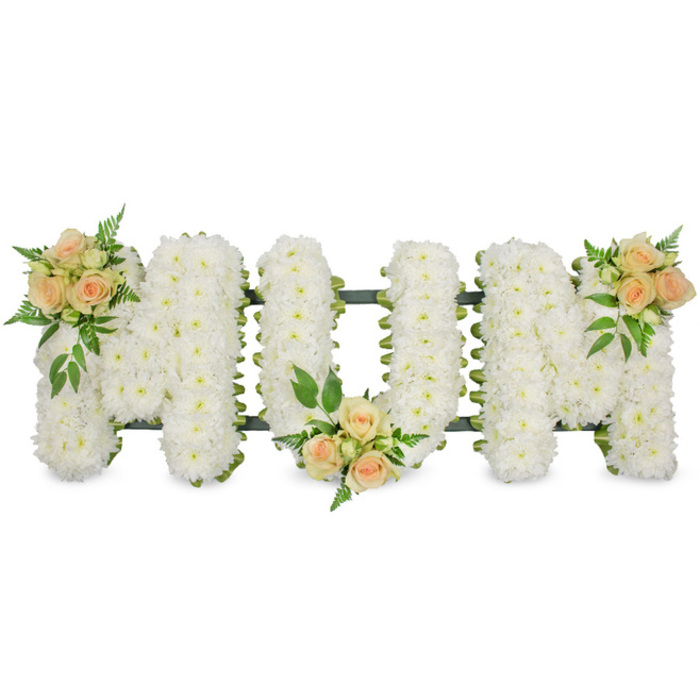 This long, horizontal floral arrangement clearly spells out MOM in three substantial capital letters, carefully arranged for a funeral, cremation, or memorial service. The letters are constructed on a sturdy frame and then completely covered with countless white spray chrysanthemums, each bloom small, rounded, and immaculate. Pressed closely together, they form a thick, cushion-like layer of petals, creating a uniform, snowy surface that appears both soft and serene. The edges of the letters are lightly defined by a narrow band of dark green ribbon and structural foliage, which helps to separate each character visually and adds a subtle contrast to the whiteness. To break up the expanse of white, three tastefully placed rose clusters introduce colour and emotional nuance. These accents feature soft peach and apricot-hued roses, touched with blush pink tones, some fully open to reveal their layered centres and others still held tightly in bud. The first cluster decorates the upper left of the initial M, the second nestles along the bottom of the central O, and the third rests on the upper right of the final M. Each grouping is accompanied by lush greenery, including sprigs of fern and deep green leaves that peek out among the roses and chrysanthemums, lending dimension and a natural, garden-fresh feel. The tribute feels both classic and deeply personal-a floral expression of gratitude, love, and farewell for a mother. It is easy to imagine this arrangement prepared by Grove Park Florist for a local family, then delivered with care to a service near Grove Park station or by the peaceful greenery of Grove Park itself, where its quiet beauty can offer comfort to those gathered.