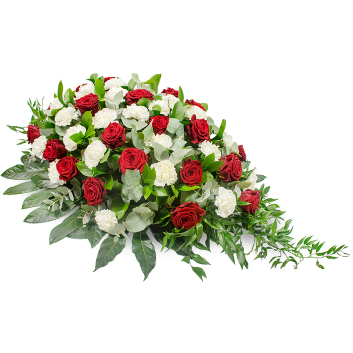 A solemn yet beautiful funeral floral arrangement is displayed on a clean white surface, its elongated, asymmetrical form drawing the eye from a fuller left side down to a refined, trailing right tip. The centre of the design is filled with deep crimson red roses and white carnations, a timeless colour pairing that carries strong emotional resonance. Each rose is tightly furled, its velvety petals forming rich, rounded blooms that sit proudly above the greenery, like individual tokens of love and gratitude. Interspersed among them, the white carnations present layers of gently ruffled petals, their bright whiteness and cushioned shapes softening the overall look and evoking innocence, remembrance, and peace. Supporting this floral duet is a substantial base of foliage. Broad, dark green leaves create a stable, glossy underlay, helping to define the outline of the spray and giving it a polished, professional finish. Running through the design, soft-toned eucalyptus in muted silvery-green adds depth and subtle variation, its slender stems and rounded leaves providing an airy, slightly textured contrast. At the far right, slender trails of fine greenery extend beyond the main body of flowers, offering a flowing, almost river-like movement that brings to mind the gentle curves of the Thames not far from Grove Park. The lighting in the image is even and bright, leaving no harsh shadows, so every petal, leaf, and edge is clearly visible. This is the sort of dignified tribute that could rest with quiet grace on a casket in a Grove Park church or be placed near the front of a crematorium chapel, providing a focal point for reflection and farewell.