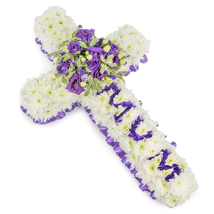 A substantial, reverent cross fashioned from an expanse of closely arranged white chrysanthemum blooms, forming a plush, pillowy field that reads as serene and consoling. The chrysanthemums are chosen and placed with precision so the surface appears uniform and almost tactile, the petals creating a soft matte texture that reflects a subtle, powdery aroma. Encircling the perimeter is a ruffled border of purple satin ribbon, its pleats catching the light and lending a dignified frame to the white base. Running down the vertical beam, the word MUM is assembled in bold purple florets - smaller eustoma or spray chrysanthemums - their deep tones from lavender to violet giving the dedication a graceful prominence. Anchoring the composition at the cross's centre is a lush floral spray: concentrated violet and lavender roses with compact buds, airy lisianthus with ruffled edges, and pale eustoma layered against variegated greenery, possibly pittosporum or eucalyptus, whose light-edged leaves and darker centres add depth; tiny green berries are threaded through to lift the texture. The piece is set against a clean white backdrop and lit with soft daylight so that the cool whites and layered purples register with calm clarity. This design is particularly suited to funeral services in the Grove Park and Lewisham area, whether placed in a local church, chapel, or brought to graveside in nearby cemeteries; it conveys a composed mix of respect, sorrow, and affectionate remembrance, a timeless floral gesture from neighbourhood florists familiar with the local community.