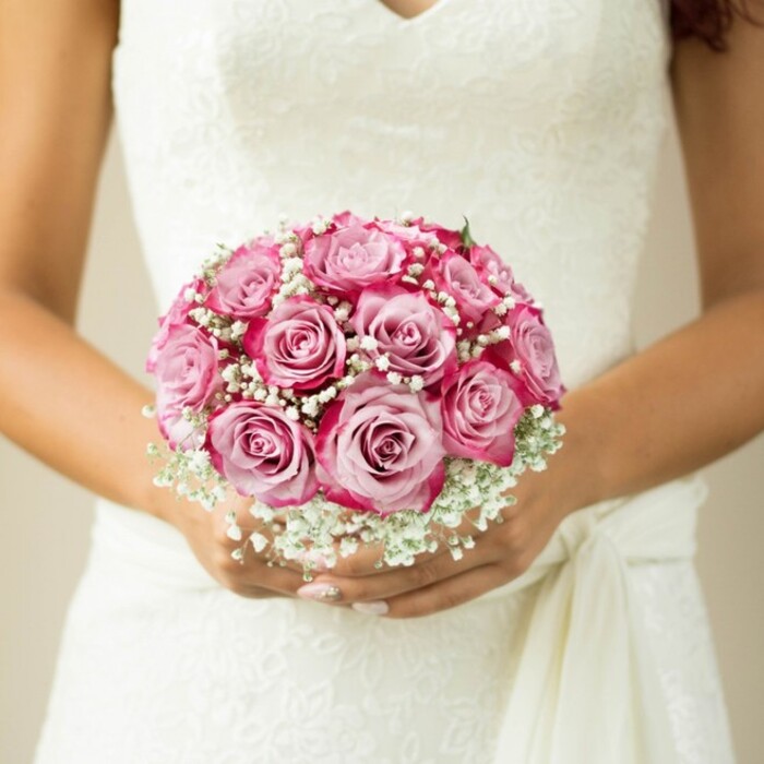 This evocative close-up, set against the refined simplicity of a Grove Park backdrop, tells the story of a bride treasuring a lush, round bouquet. Her hands, resting lightly atop the elegantly detailed bodice of her white wedding gown, cradle a composition dominated by roses in perfected bloom. Deep magenta tones radiate from the edges of each rose's velvety petals, fading delicately to blush-pink at their core-the sort of transition seen in the finest floral arrangements across Grove Park's high streets. Misty sprays of baby's breath weave softly between the roses, imparting a cloud-like softness that enhances the bouquet's visual and emotional depth. Only a touch of green foliage peeks through, hinting at the bouquet's tightly gathered stems. The dress-sleeveless, floral, and detailed with lacework over the V-neck-embodies the spirit of elegant celebration, with a sash near her hands finishing the look. The light, neutral blur of the background holds the composition together, ensuring all eyes are drawn to the artistry of the bouquet and to the poignant, joyful anticipation that defines wedding days prepared by florists in Grove Park.
