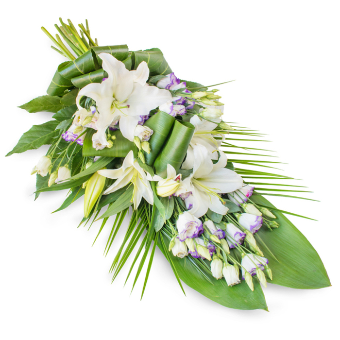 This image shows a sophisticated memorial spray set diagonally on a plain white background, with the body of the arrangement gathered fully at the upper left and trailing in a slender, pointed line towards the lower right. Dominating the design are several white oriental lilies, some open into wide, expressive blooms with curved, slightly ruffled petals and visible yellow-green stamens, others standing as elegant, tapered buds. These lilies provide a focal sense of purity and light. Around and between them, the florist has woven clusters of Lisianthus-smaller blooms shaped like soft bells, their white petals trimmed with a delicate wash of lavender or pale purple, with fine veins that could be felt more than seen. Numerous Lisianthus buds introduce small, rounded forms that play against the longer lines of the lilies. Beneath and surrounding the flowers is a layered foundation of foliage. Broad, dark green leaves form a quiet, supportive background, while certain leaves are rolled and folded into structured loops that add a modern, almost sculptural flair near the upper centre. From the right-hand side, slender, palm-like fronds fan outward, slightly arched, echoing the natural sweep of branches along Grove Park's tree-lined streets and reinforcing the elongated, flowing silhouette. The overall effect is calm and poised, primarily white and green with that soft whisper of lavender, ideal for a funeral service or cremation gathering in Grove Park or nearby churches. Up close, one might notice the cool, waxy feel of the leaves and the subtle fragrance of lilies and fresh greenery, a restrained scent suited to quiet, reflective spaces. The spray communicates respect, remembrance, and a gentle, enduring love through its balanced composition.