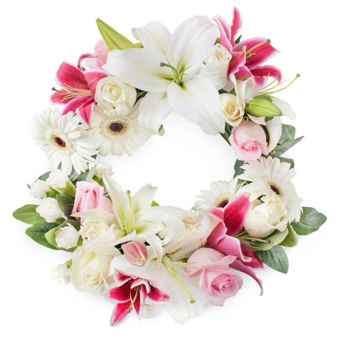 A beautifully detailed funeral wreath fills the frame, its circular, open-centre shape resting against a simple white background that lets the flowers speak quietly for themselves. The design is opulent yet restrained, featuring a careful blend of white, blush, and deep pink blooms that together create a calm but emotionally resonant tribute. Large white lilies are the stars of the arrangement, their star-shaped petals fully open in places, with some blooms still in gentle stages of unfolding. Their creamy, luminous surfaces and prominent golden stamens bring a sense of purity and light. Threaded carefully amongst them are striking deep magenta and fuchsia lilies, their richer tones and speckled markings adding a layer of depth and intensity, like treasured memories that remain vivid even in sorrow. Around the lilies lie clusters of light pink roses, some tightly furled, others partially open, the softness of their velvety petals evoking affection and remembered kindness. Full, creamy-white roses provide classical structure and volume, their layered petals nestled slightly lower in the wreath, creating a pleasing sense of depth. Bright white gerbera daisies punctuate the ring, their dark central discs and neat, evenly spaced petals adding rhythm and a contemporary feel. Small white filler flowers-delicate blooms reminiscent of miniature carnations or spray chrysanthemums-fill the spaces between larger blossoms, ensuring the wreath appears full and harmonious from every angle. A collar of fresh green foliage frames the floral ring, with leaves of varying sizes and shades overlapping to create a natural, garden-like effect. It is easy to imagine this wreath prepared by Grove Park Florist and delivered to a quiet service near Grove Park station or a peaceful chapel along the Thames, offering a gentle, fragrant expression of condolence and respect.