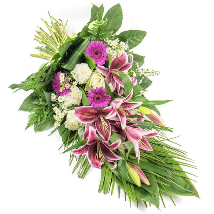 A beautifully elongated funeral sheaf is set against a spotless white background, highlighting its refined shape and harmonious palette. The arrangement flows diagonally from the top left, where the stems are firmly bound together, down to a tapered point on the lower right, forming an elegant crescent or teardrop silhouette. At the heart of the sheaf, large Stargazer lilies in rich magenta and fuchsia tones open wide, their petals edged with white and speckled with burgundy, some fully outstretched, others slowly unfurling or still in bud. These striking blooms are gently surrounded by clusters of white roses with creamy centres, their petals tightly furled or just beginning to open, expressing purity, reverence and quiet devotion. Ruffled white carnations add depth and softness, cushioning the stronger shapes of the lilies and roses. Punctuating the design, vivid magenta gerbera daisies stand out, their flat, daisy-like faces adding a modern, heartfelt brightness. Interspersed throughout are tiny white filler flowers, similar to miniature daisies or gypsophila, whose delicate blossoms lend a lace-like lightness. Enveloping the flowers, a generous collection of green foliage provides structure and movement: broad, dark green leaves form a solid base, while feathery fern fronds soften the outline and long, slender, grass-like leaves arc outward and downward to emphasise the spray's flowing curve. The lighting is even and soft, revealing the satiny sheen of the lily petals and the velvety texture of the carnations. This tribute feels like a piece created by an experienced Grove Park florist for a service in a nearby church or for delivery to a family home along quiet residential streets, a serene and respectful way to say a final, loving goodbye.