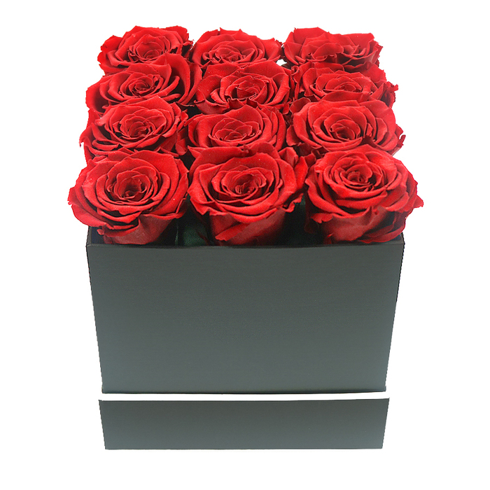 This image offers a close, contemplative view of a modern rose box arrangement, evoking the refined charm of Grove Park's riverside homes and leafy avenues. At its centre sits a perfectly square, matte black box, the finish smooth and unadorned, with sharp, clean edges that give it an architectural presence. The only decorative detail is a fine, bright white stripe that circles the lower portion of the box like a minimalist trim, echoing the crisp lines of contemporary interiors around Chiswick and Grove Park. Emerging from the open top are twelve deep red roses arranged with deliberate precision in a three-by-four grid. Each bloom is fully open yet beautifully intact, petals layered tightly in graceful spirals that create a velvety, almost plush surface. The red hue is intense and romantic, with slight shifts in tone where the soft light catches the petal edges, suggesting high-quality preserved roses selected for their consistency and longevity. The blooms sit closely together, their heads just surpassing the rim of the box, so that the arrangement appears both tightly composed and luxuriously full. The background is a flawless white, devoid of any other elements, allowing the eye to rest solely on the interplay between the rich roses and the dark, square container. Gentle, even lighting casts only the faintest of shadows, enhancing the texture of each petal and the crisp silhouette of the box without introducing harsh contrasts. The atmosphere is one of poised romance and quiet indulgence, the kind of floral gift you might imagine delivered same-day to a Grove Park address for an anniversary, a special dinner overlooking the river, or a thoughtful gesture that expresses deep affection in a subtle, elegant way.