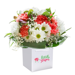This garden-fresh bouquet, carefully composed by local Grove Park artisans, radiates an inviting blend of colour, fragrance, and texture. Nestled upright in a crisp white gift bag, the arrangement hums with the lively interplay of lush coral and peach carnations-their layered, ruffled petals seem to ripple in the gentle light-balanced by snowy daisies crowned with bright greenish-yellow centers. Baby's breath, airy and light as dandelion fluff in Chiswick Park, weaves delicately among the larger blooms, filling the gaps with soft poise and a whisper of nostalgia. The occasional aubergine-tinted bud on the left adds intrigue, hinting at late spring promises yet to unfold in local gardens. Deep, glossy leaves fan out around the base, creating a generous verdant framework that holds the flowers aloft, much like the sheltering trees along Sutton Court Road. The whole arrangement is designed for vibrant presence and effortless gifting, with sturdy ribbon handles securing the white bag. Isolated against a snowy backdrop, each detail is accentuated-each carnation, daisy, and flourish of greenery a testament to the pride and care Grove Park Florist pours into its work. Ideal for marking milestones, sending comfort, or simply brightening a friend's kitchen table near the Thames, this stylish bouquet brings both warmth and sophistication to every Grove Park doorstep.