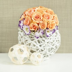 On a clean, reflective white tabletop that catches gentle light, a carefully considered floral arrangement becomes the focal point, much like a centerpiece set for a wedding breakfast or engagement dinner in Grove Park. The upper portion of the design is a lush, rounded dome of roses in varying peach and apricot hues, each bloom fully open and cushioned close against its neighbour, so that the surface appears almost like one continuous sweep of silky petals. The roses' colour shifts subtly from pale peach at the crown to warmer apricot tones as the eye travels downward, adding depth and richness. Cradling the base of this rose dome is a ring of white lisianthus, their petals gracefully fluted and finely edged in rich violet and purple, providing a striking yet refined contrast. Interspersed are small sprigs of white baby's breath, their delicate clusters like tiny clouds, filling the spaces between the lisianthus and enhancing the softness of the design. All of the blooms are arranged in a distinctive white basket-style vase, broadly spherical and crafted from interwoven wicker strands that form a bulbous open lattice, revealing glimpses of interior shadows. In front of the vase, two decorative woven balls made from light, natural wood-toned strips lie on the same white surface; their airy pattern echoes the vase's texture and offers a playful, modern touch. Behind the arrangement, a softly textured beige background resembling fine fabric or wallpaper provides a warm, understated setting. The entire scene suggests romance, calm, and quiet sophistication, fitting for wedding flowers prepared by a local Grove Park florist for celebrations at home or nearby venues along the Thames.