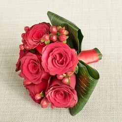A compact and luxurious bouquet of red roses and berries is carefully arranged on a soft, light beige fabric with a subtle woven texture, like linen laid out on a polished table in a Grove Park townhouse. At the heart of the design are several large, fully opened red roses, their petals velvety and lush, blending deep crimson with slightly lighter scarlet highlights where the light gently brushes their curves. The roses are tightly grouped, creating a romantic focal point that feels both classic and modern. Threaded through the blooms are clusters of small, round Hypericum berries in shades of reddish-pink and berry-red, their smooth, bead-like surfaces catching the light and introducing a playful contrast to the softness of the petals. Dark green tropical leaves, broad and structured, have been expertly curled and folded around the base, forming an elegant collar that supports and frames the flowers. A rich red satin ribbon is wrapped firmly around the stems, its sheen adding a final touch of sophistication, reminiscent of a bridesmaid bouquet carried down the aisle at a wedding near Grove Park station or along the quiet paths toward Chinbrook Meadows. The lighting in the image is even and calm, with no harsh highlights, allowing the eye to linger on the luxurious textures and saturated colours. This arrangement feels perfectly suited to romantic occasions, from intimate proposals and Valentine's surprises to special dinners celebrated at home, embodying the careful, personal touch you would expect from an experienced local florist familiar with the rhythm of life in Grove Park.