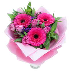 This composition reads like a gentle love letter from a local florist in Grove Park, designed to lift spirits and mark an occasion with warmth. At its heart are three fuchsia-pink Gerbera daisies, each face open and exuberant, their radiating petals surrounding a distinct dark centre that adds bold contrast and visual focus. Complementing them are several roses in a soft lavender-pink or dusty-rose tone, their petals tightly rolled as when newly cut, giving the bouquet a tender, budding quality. Interspersed are clusters of vivid purple statice, which float between the main flowers and introduce delicate texture and a cool, duskier hue. A rich patch of greenery - broad leaves that read as salal or eucalyptus - creates a verdant backdrop, the glossy surfaces catching light and emitting a faint, fresh scent. The flowers are cocooned in an inner layer of pale pink tissue paper and an outer sheet of translucent, crinkled fuchsia-pink cellophane that catches glints of daylight; the wrapping is folded with considered hands, ready for handover or same-day delivery across Grove Park. The top rim of a clear glass vase peeks out beneath the wrapping, suggesting the bouquet is arranged for immediate display on a mantle or kitchen table. The overall feeling is celebratory yet intimate: a present fit for birthdays, anniversaries, Mother's Day or a simple 'thinking of you' moment. Crafted with local care by Grove Park Florist, this bouquet balances cheerful colour, soft fragrance and thoughtful presentation to make the recipient feel genuinely noticed.
