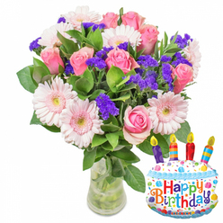 A serene, joyful bouquet is captured here: the arrangement sits against a crisp white background and reads like a small garden harvested at morning's light-soft pink roses that move from a whisper of blush to richer pink cores, surrounded by pale-pink Gerbera daisies whose broad petals open like warm faces. The roses' layered petals feel velvety to the touch, while the Gerberas bring a lighter, almost porcelain brightness. Interspersed throughout are compact clusters of vivid purple filler flowers-probably statice or limonium-whose feathery, papery clusters introduce a contrasting texture that makes the pinks sing. Little white spray blooms, perhaps tiny carnations or alstroemeria, add delicate accents, and a generous sweep of glossy green foliage gives the bouquet a grounded, lush silhouette. The stems are visible through the clear glass vase, the water catching subtle reflections and confirming the flowers' freshness.

Beside the vase, a cheerful cake-shaped metallic foil balloon amplifies the celebratory mood: a frosted-white design trimmed with sky-blue piping, colorful sprinkles in red, yellow, green and blue, and four colorful candles-purple, red, orange and blue-each with a painted yellow flame. Happy Birthday is printed in bright, playful letters across the cake, and a small KALEIDOSCOPE mark rests at the bottom edge. The composition feels like an offered moment of celebration-softly scented, texturally rich, and composed with the thoughtful hand of a florist attuned to feeling as much as form.