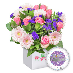 The bouquet arrives as a sunlit memory of spring, a joyful statement lovingly crafted for a recently engaged couple. A classic pairing of soft pink roses, their petals unfurling in gentle spirals from tight buds to open blooms, fills the centre with whispered romance, while pastel pink gerbera daisies radiate warmth, their white edges catching the light and revealing pale yellow anchors at their hearts. Between these starry blooms, clusters of vivid purple filler flowers peek out, lending velvet depth and a dash of playful contrast to the pastel sea. A veil of lush green foliage flows around and between the stems, providing a garden-fresh backdrop that makes the colours feel truly alive. All of this rests in a clean white square box that offers a modern stage for the flowers to shine, the front bearing a discreet red rose emblem that hints at craftsmanship and care without shouting. A round foil balloon-partially visible on the lower right-carries the triumphant message 'You're Engaged!' scripted in shimmering silver, with a central purple circle setting a formal highlight for the celebration. The little golden ring motif completing the O adds a subtle spark of symbolism, while the surrounding tiny hearts and silver stars embellish the air with romance. Delivered straight to homes or offices across Grove Park and nearby neighbourhoods, this bouquet is more than flowers; it's a note of happiness, a promise of new beginnings, and a gesture that makes a celebratory moment feel warmly personal for the couple.