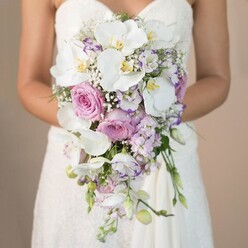 A soft haze of morning light caresses a bride's hands as she cradles a grand, cascading bouquet-its gentle drama echoing the understated charm of Grove Park. The bouquet's form is unmistakably elegant: broad white phalaenopsis orchids gleam like pearls, their luminous petals contrasting against blushing pink garden roses. Between these, clouds of delicate lavender-toned stock and frilled lisianthus unfurl, their subtle purple hues reminiscent of springtime strolls through Dukes Meadows or the residential streets nearby. Sprigs of airy baby's breath and wisps of fresh greenery lend the arrangement a feathery, organic movement. The bride's arms, adorned in a strapless gown with intricate lace, direct attention to the bouquet's sweeping lines and textured clusters. Every petal and stem reveals the attention to detail and artistry that Grove Park Florist brings to local weddings. The bouquet emanates not just visual beauty, but the sense of anticipation, tenderness, and celebration unique to Grove Park's most cherished occasions-a perfect choice for couples seeking both timeless tradition and contemporary floral style.