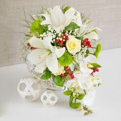 On a clean white tabletop, an elegant floral arrangement is displayed as though prepared for a wedding breakfast in Grove Park, with a soft beige, lightly textured background that sets a serene, neutral stage. The flowers are arranged in a white woven wicker basket, the basket's subtle criss-cross pattern complementing the organic lines of the blooms. Dominating the composition are several large white lilies, wide open and trumpet-shaped, their creamy petals lightly speckled and their stamens tipped with warm pollen that suggests a sweet, heady perfume. Nestled close to the upper center of the arrangement is a single cream-yellow rose, its tightly layered petals forming a romantic focal point that draws the eye inward. To the right, a graceful trail of white phalaenopsis orchids cascades downwards, each bloom like a delicate butterfly alighting on the stem, creating a soft waterfall effect that brings movement and elegance. Around these main flowers, tufts of bright green pom-pom chrysanthemums add playful texture and a fresh pop of colour, mingling with fine, feathery greens and broader, glossy leaves that evoke garden foliage from nearby Chiswick or the Thames path. Tiny white baby's breath blossoms thread through the arrangement, softening transitions between the larger blooms, while clusters of glossy red berries offer striking accents, catching the light and lending a festive undertone. On the left side of the scene, two white wicker spheres, one larger and one smaller, rest on the table, their light, open construction mirroring the basket and adding a modern decorative flourish. The entire display feels luxurious yet inviting, a sophisticated centerpiece or bridal table arrangement ideal for intimate celebrations and same-day flower delivery throughout Grove Park.