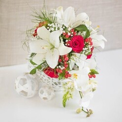 An elegant and romantic floral arrangement sits as a focal point on a white tabletop, lit by soft, even light that gives the scene an intimate, serene character, much like a quiet wedding reception in Grove Park. The flowers are gathered in a round, white open-weave basket, its delicate latticework offering glimpses of stems and creating a light, contemporary feel. Out of this airy vessel, several large white Oriental lilies extend, their broad petals opening wide to reveal gently arched stamens tipped with golden-brown pollen. The lilies' cool, creamy white surfaces contrast beautifully with the rich tones of the surrounding red roses, whose velvety petals are tightly clustered into lush, romantic blooms. Throughout the design, sprigs of fine white baby's breath introduce a soft, mist-like texture, while slender bright green foliage and feathery greenery lend freshness and subtle movement, as if a light breeze were passing across the table. Scattered snugly among the leaves, glossy red hypericum berries appear as tiny, polished beads of colour that hint at festive dinners along Burlington Lane or cosy winter celebrations at home. On the right edge of the arrangement, a cascade of white phalaenopsis orchids drapes gracefully, the blooms spaced neatly along the arching stem, each flower revealing a delicate yellow and blush centre framed by smooth white petals. A few unopened buds trail towards the tip, adding a gentle sense of unfolding time. On the left side of the composition, three decorative white spheres, made from the same woven material as the container, rest in a loose cluster, echoing the basket's texture and balancing the scene. This refined combination of lilies, roses, orchids, and berries creates a centerpiece ideal for weddings, anniversaries, or intimate gatherings around Grove Park and its surrounding neighbourhoods.