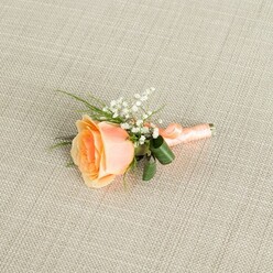 A softly glowing peach rose boutonniere is displayed on a pale beige, linen-textured fabric that resembles a finely woven table runner in a Grove Park townhouse. The fabric's subtle cross-hatch pattern stays understated, allowing the eye to linger on the single rose placed just off-centre, resting horizontally as if waiting to be pinned before a wedding ceremony. The rose itself is partially opened, its petals unfurling in gentle curves from a richer apricot heart to light, creamy outer edges, creating the impression of warm dawn light captured in a flower. Nestled against the upper side of the bloom are feathery sprays of white baby's breath, each minuscule blossom adding sparkle and a sense of lightness. Slender strands of green foliage, along with two broader dark green leaves that arch protectively around the base, lend depth and a polished outline to the composition. The stems are bundled tightly and wrapped in a smooth peach satin ribbon, wound with the kind of care a professional florist from Grove Park Florist would take before sending it out for same-day delivery across the neighbourhood. The ribbon ends in a neat, tidy finish that sits snug against the stems, suggesting comfort and ease when worn on a jacket lapel. Gentle, even light falls across the scene, revealing the velvety texture of the rose petals, the delicate clusters of gypsophila, and the slight sheen of the ribbon. The overall mood is one of quiet, romantic anticipation, as if this boutonniere were prepared for a groom waiting at a local Grove Park church or registry office, ready to mark a cherished new beginning.