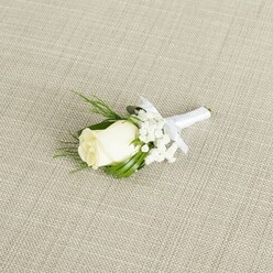 A refined wedding boutonniere rests on a pale beige, linen-like fabric, the subtle crosshatch of the weave evoking the understated elegance of a Grove Park sitting room prepared for a special occasion. The focal point is a single creamy white rosebud, still tightly wrapped, its smooth, velvety petals arranged in a graceful spiral that draws the eye inward. Just at the base of the bloom, faint hints of soft green from the sepals peek through, suggesting freshness and careful handling. Surrounding the rose are airy clusters of white baby's breath, each tiny blossom forming a light, misty frame that softens the overall composition in the same way morning mist softens the view over the nearby Thames. Slender, deep emerald leaves, along with a few feathery sprigs of greenery, fan out around the flowers, giving depth, structure, and a natural rhythm to the design. The stems are firmly wrapped in a pristine white satin ribbon, its surfaces catching the light with a gentle sheen, and tied into a neat, proportional bow positioned just under the floral head, ready to be pinned to the lapel of a tailored suit. The lighting in the scene is soft and even, without glare, accentuating the gentle shadows between petals and the crisp edges of the leaves. This boutonniere conveys quiet sophistication and romantic anticipation, perfectly suited to a groom walking through Grove Park towards a ceremony in a local church or garden venue, where every detail-from the flowers to the attire-has been thoughtfully chosen to express love, commitment, and a timeless sense of style.